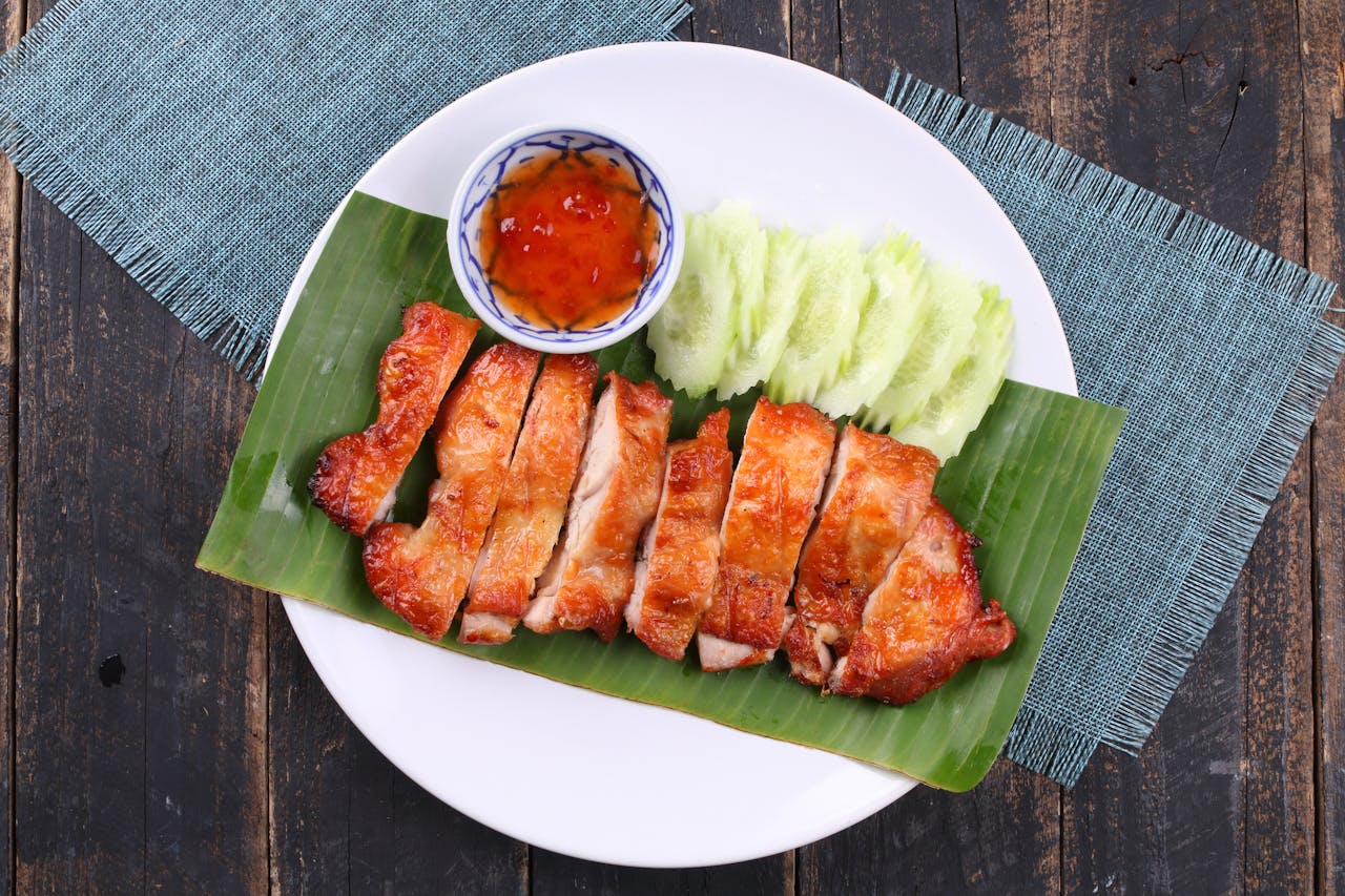 Delicious grilled chicken with spicy Thai dipping sauce and fresh cucumber served on a banana leaf.